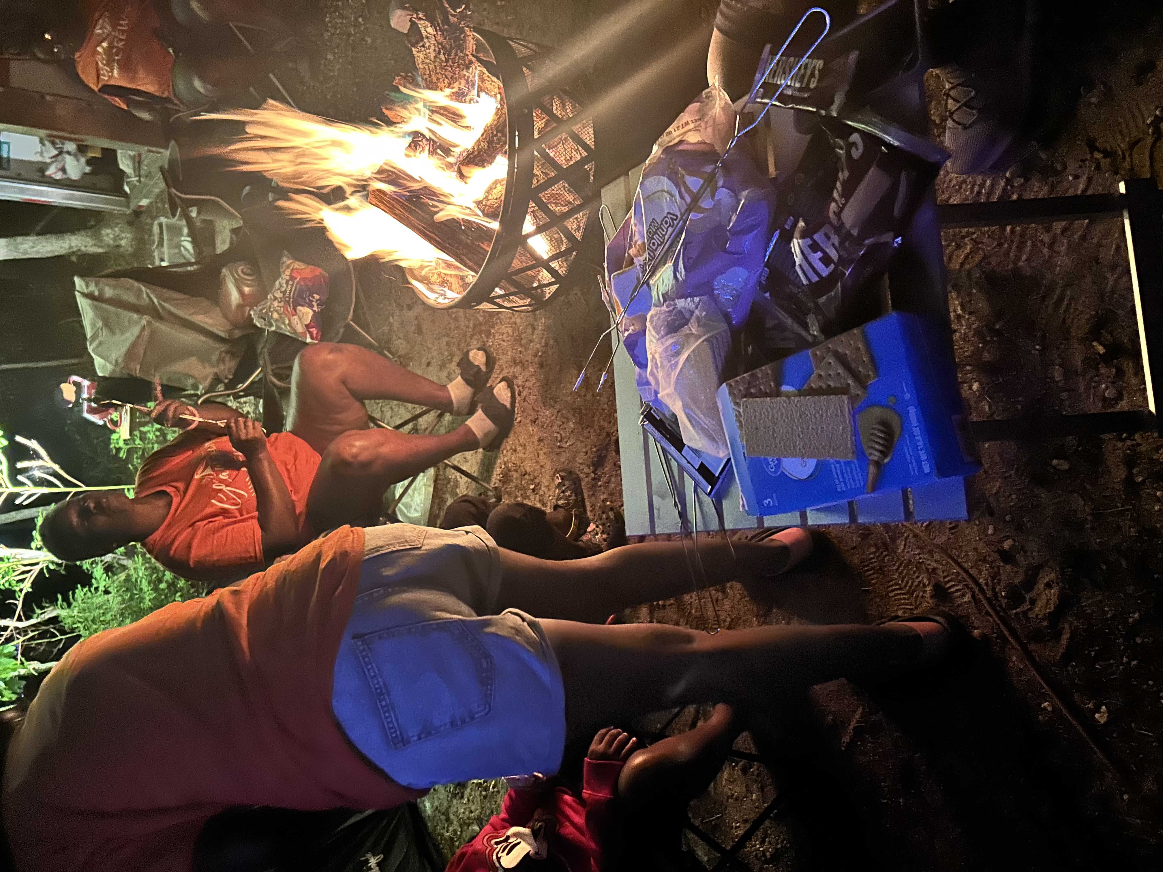 People gathered around a campfire at night, with s'mores ingredients on a table.