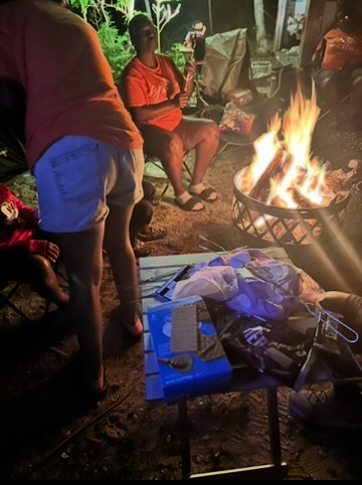 A nighttime scene around a glowing campfire, showing people gathered, with s'mores ingredients (graham crackers, marshmallows) on a small table in the foreground.