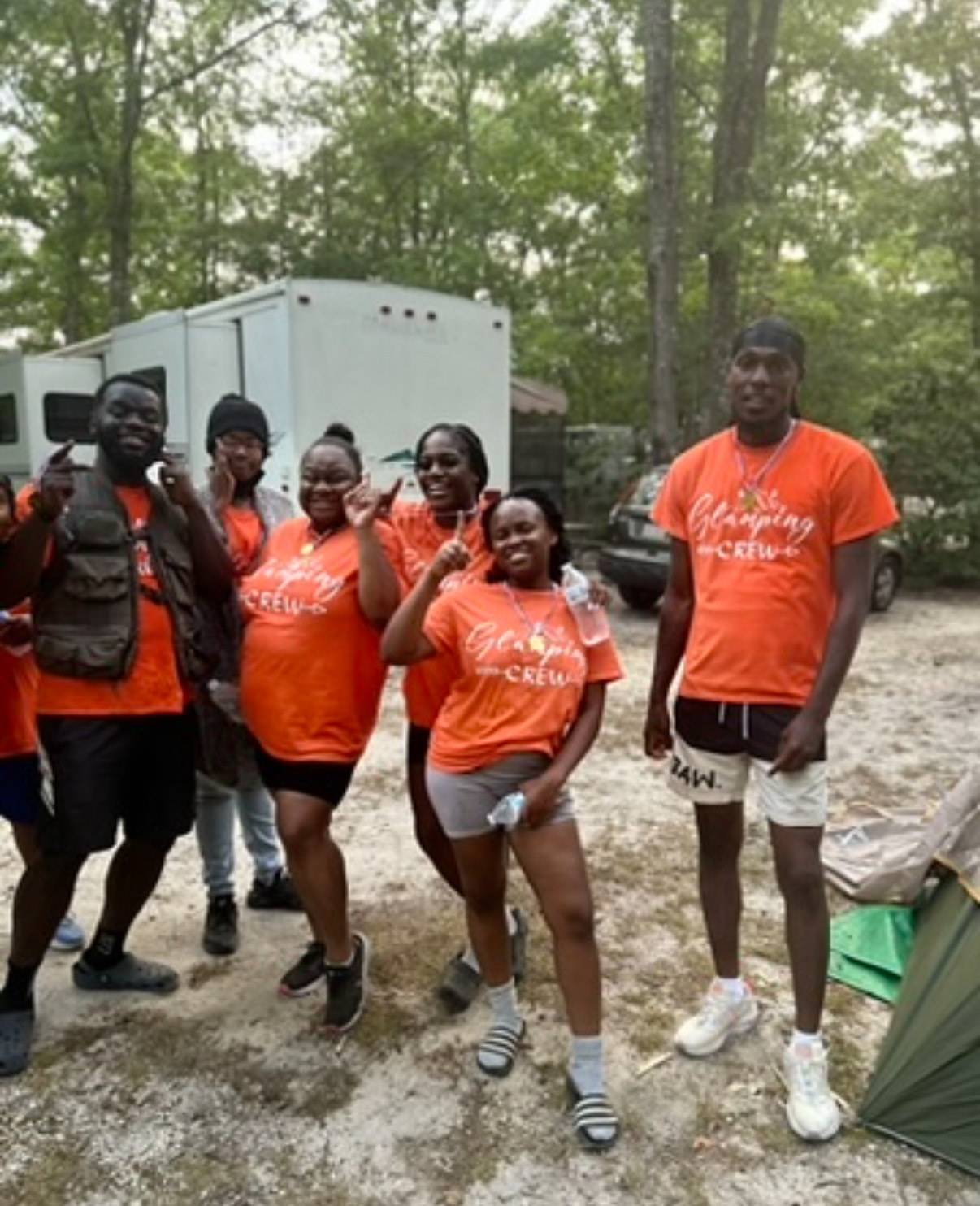 A group of adults wearing orange 'Glamping Crew' t-shirts, smiling and posing with peace signs in a wooded outdoor setting, with a white camper trailer in the background.