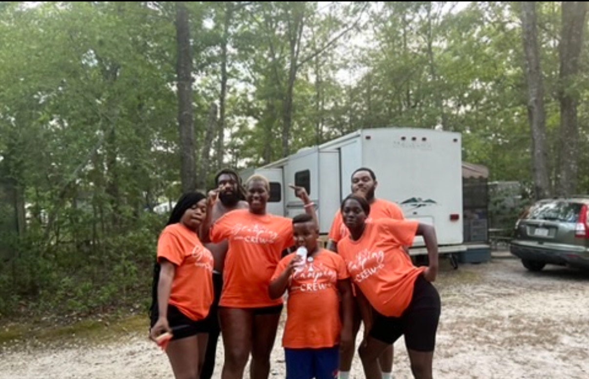 A group of adults and a child, all wearing orange 'Glamping Crew' t-shirts, posing together in a wooded area with a white camper trailer behind them.