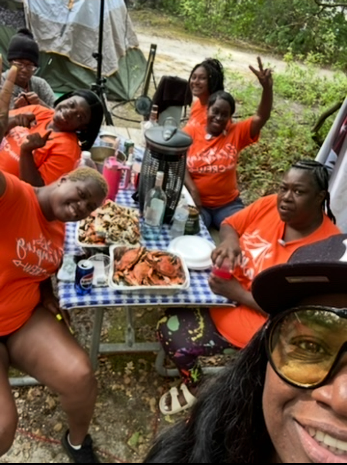 A group of people, mostly women, wearing orange 'Glamping Crew' t-shirts, gathered around a picnic table with food (including crabs) and drinks, smiling and posing for a selfie at a glamping site.