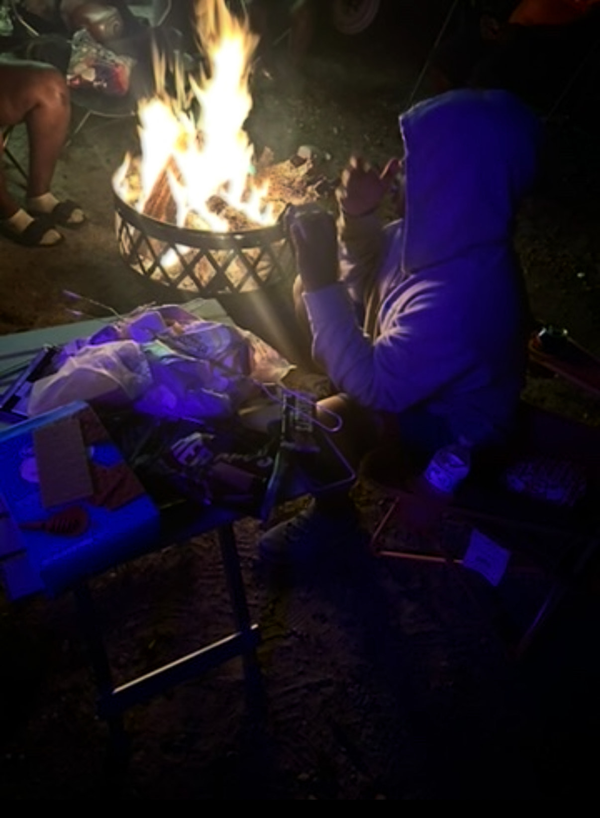 A person in a hooded sweatshirt sitting by a campfire at night, with a table holding s'mores ingredients next to them.