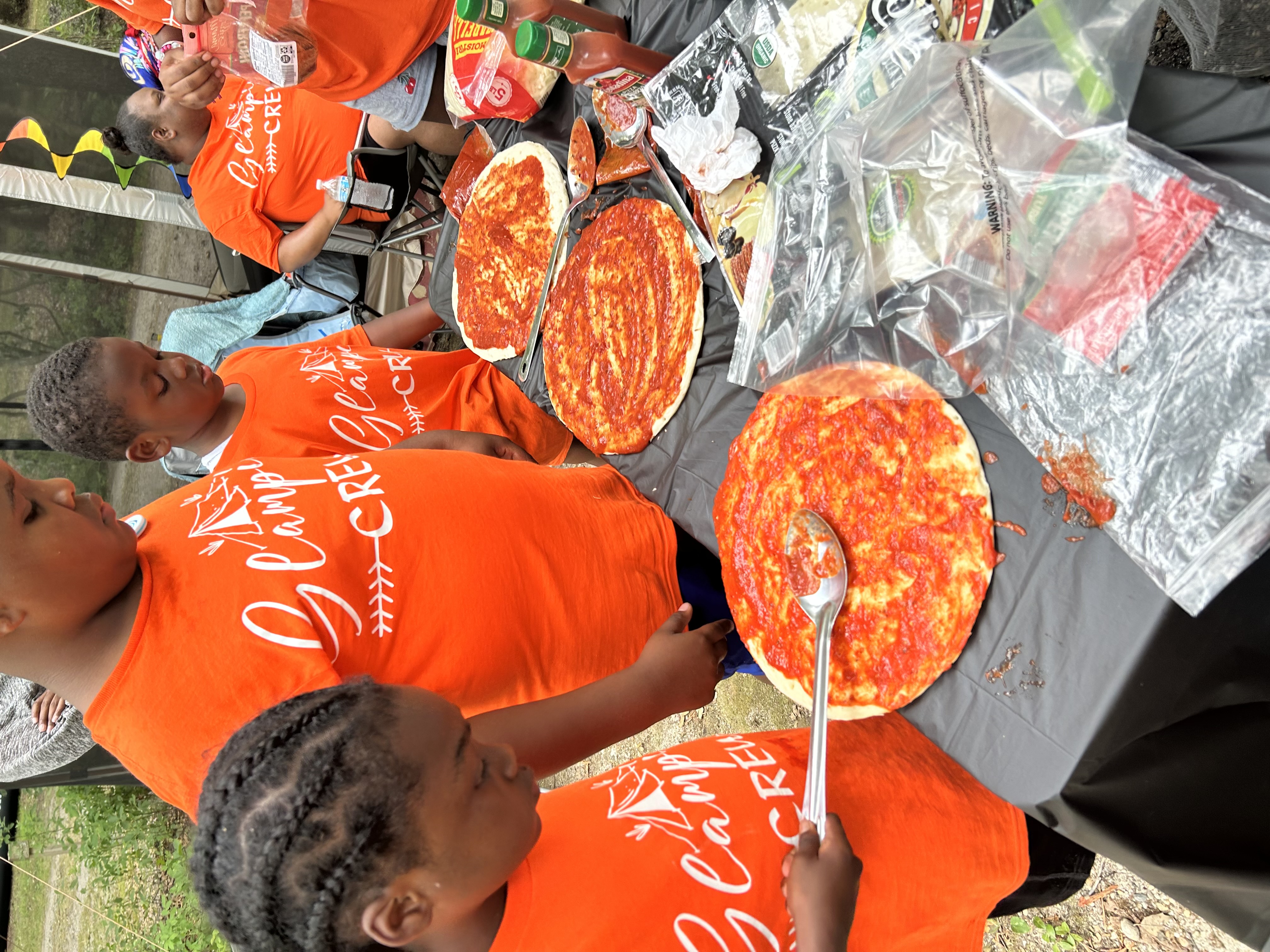 Children in orange 'Glamping Crew' shirts preparing pizzas outdoors.